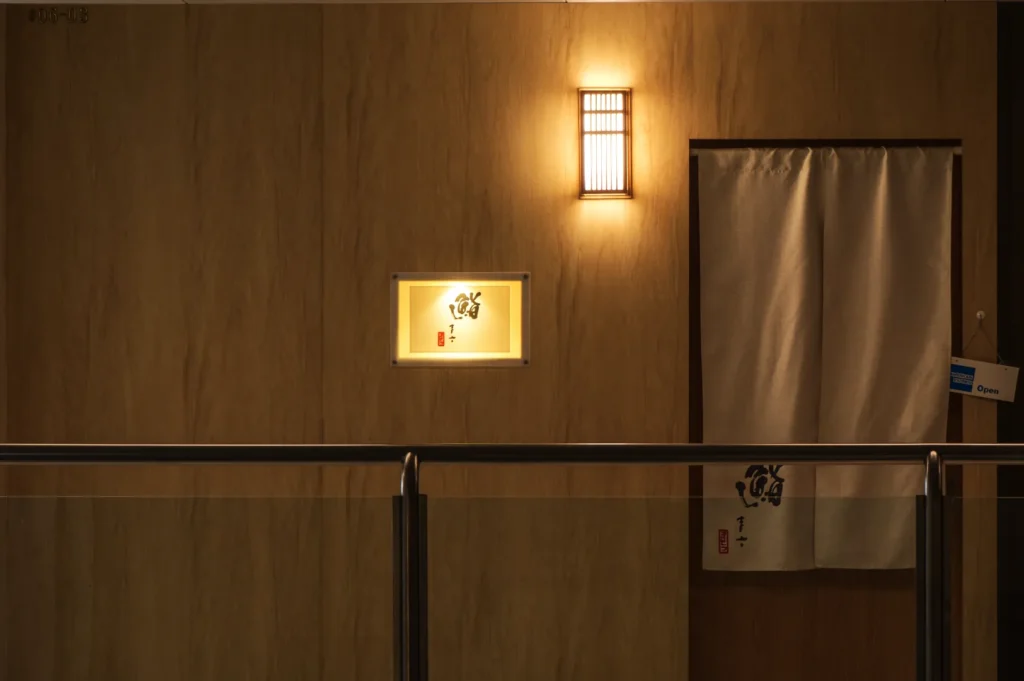 Dimly lit wooden wall with a soft glow from a Japanese-style lantern. A canvas drape hangs over a doorway, creating a serene and inviting ambiance.