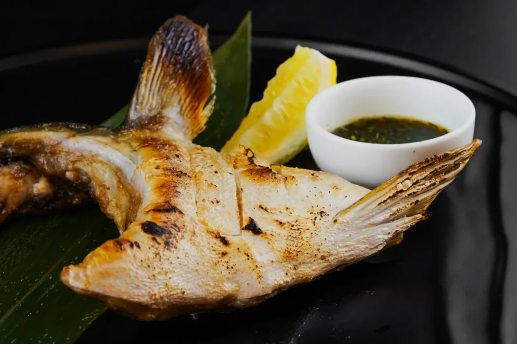Grilled fish tail on a black plate with a vibrant green leaf, lemon wedge, and a small white bowl of green dipping sauce, creating a fresh and appetizing presentation.