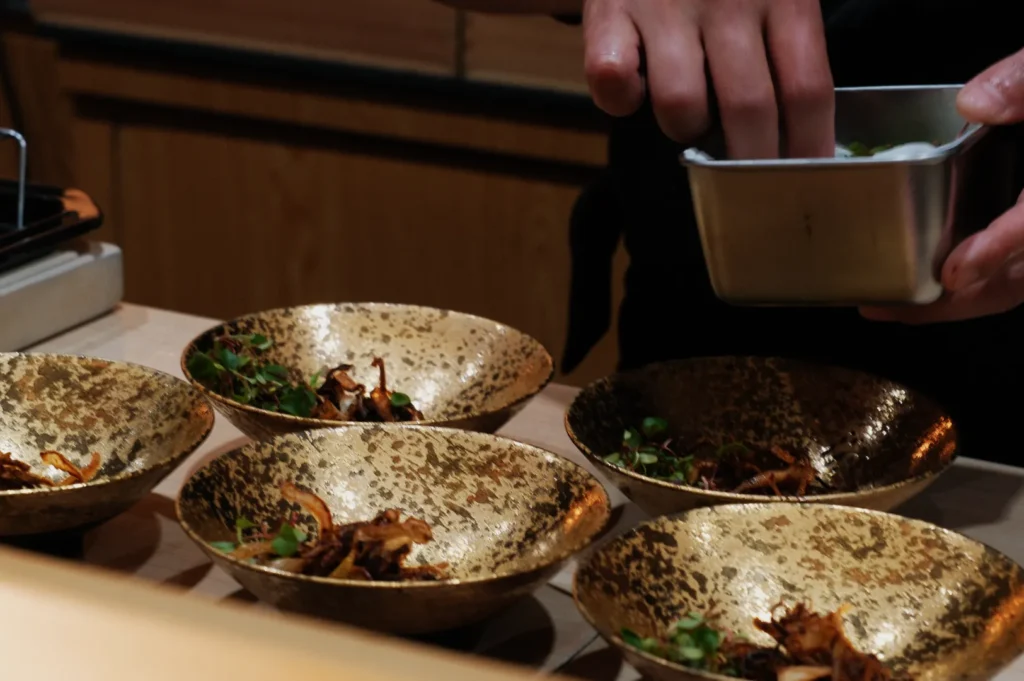 Chef garnishes elegant gold bowls with fresh greens, creating a sophisticated dish. The setting is warm and inviting, highlighting culinary artistry.