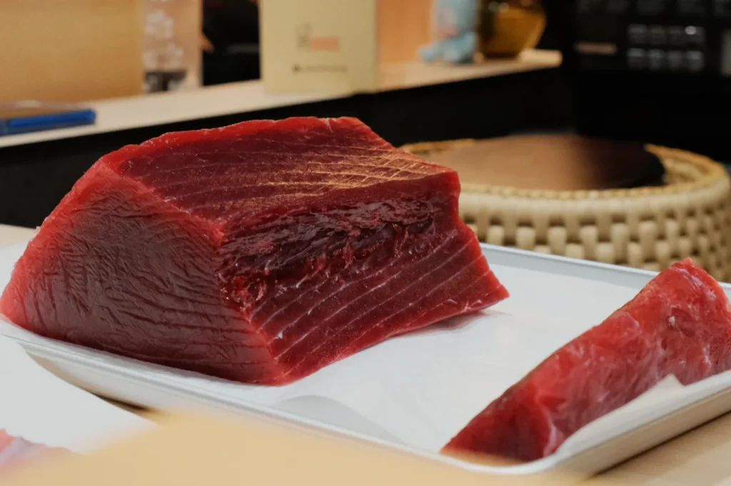 A thick, raw cut of red tuna on a white tray with a smaller slice beside it, placed on a counter in a restaurant setting, evoking freshness.