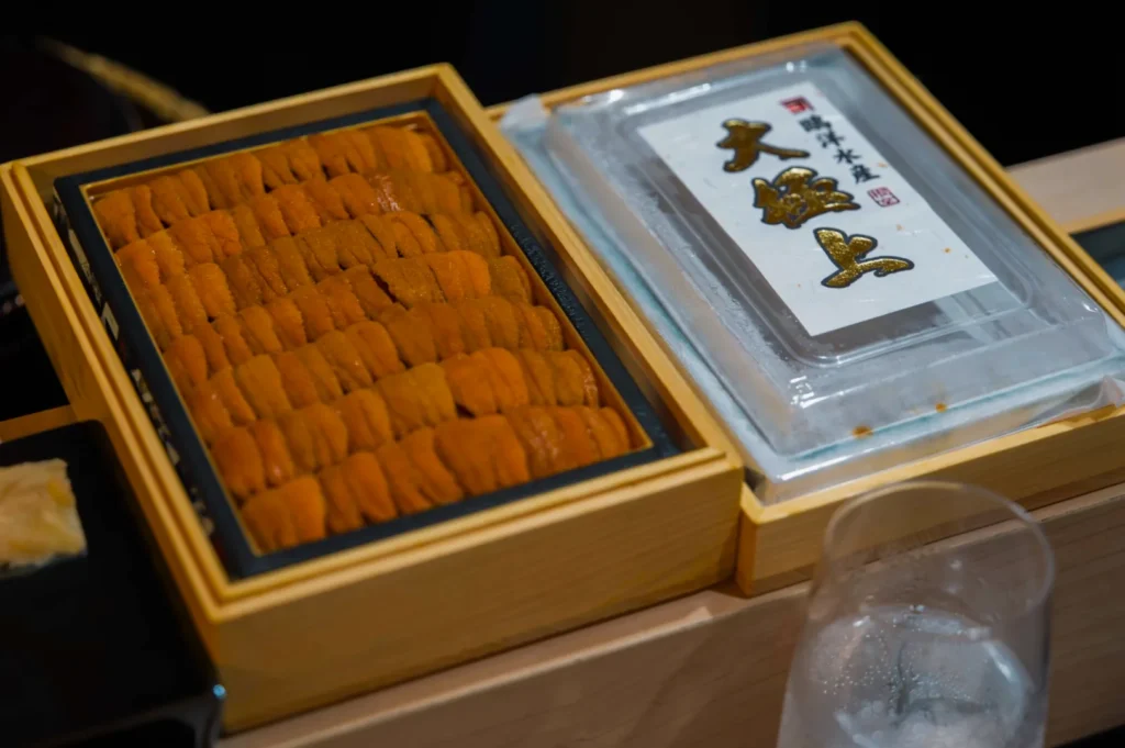 A wooden box filled with neatly arranged orange sea urchin (uni) lies next to a closed container with Japanese text. A glass with ice sits nearby.