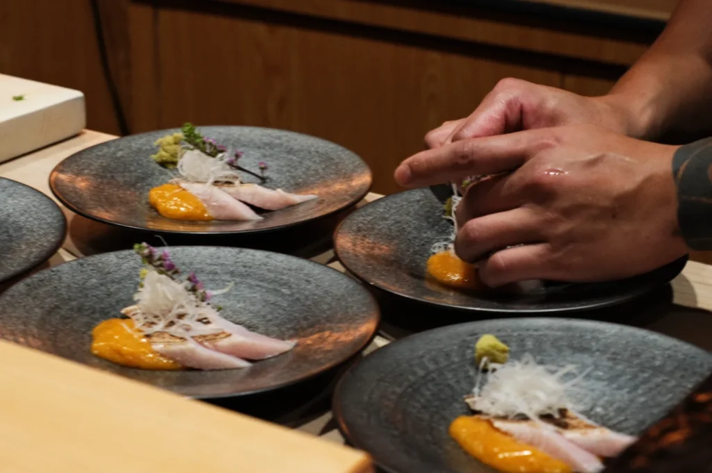 Close-up of a chef's hands plating sashimi on grey plates with dollops of orange sauce and garnished with white radish and tiny purple flowers.