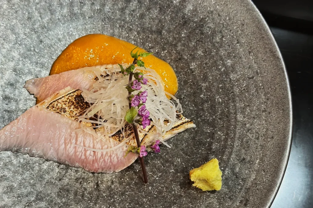Gourmet dish on a textured gray plate with seared fish, orange sauce, shredded garnish, purple flowers, and a wasabi dollop, presenting an elegant meal.