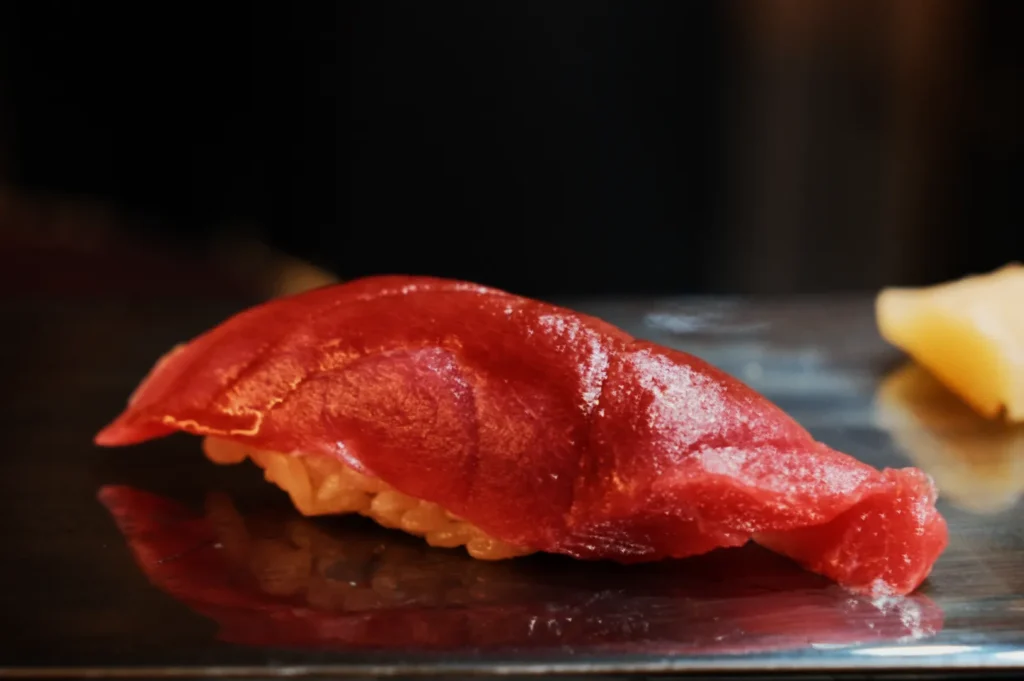 Close-up of a piece of nigiri sushi with vibrant red tuna atop white rice, set on a reflective surface. The atmosphere appears refined and appetizing.