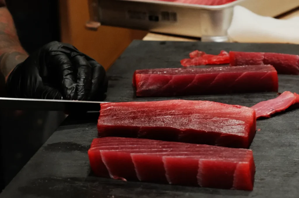 A gloved hand slices fresh tuna fillets on a dark cutting board. The vibrant red fish contrasts with the black glove, highlighting precision in preparation.