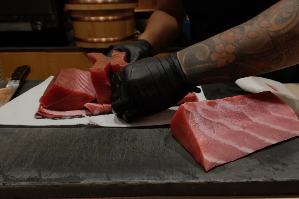 A chef with tattooed arm and black gloves slices fresh tuna on a cutting board. Thick, vibrant red tuna pieces convey precision and skill.