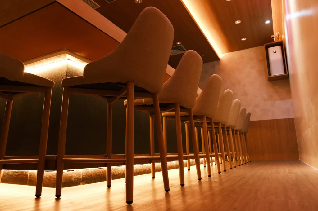 Cozy bar interior with a row of high-backed wooden bar stools. Warm ambient lighting and a modern, minimalist design create an inviting atmosphere.