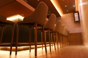 Cozy bar interior with a row of high-backed wooden bar stools. Warm ambient lighting and a modern, minimalist design create an inviting atmosphere.