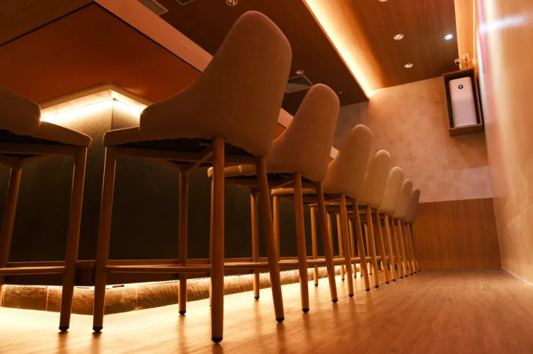Cozy bar interior with a row of high-backed wooden bar stools. Warm ambient lighting and a modern, minimalist design create an inviting atmosphere.