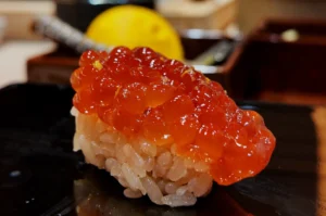 Close-up of a sushi piece with glossy salmon roe on rice, set on a dark plate. A blurred lemon in the background adds a touch of color contrast.