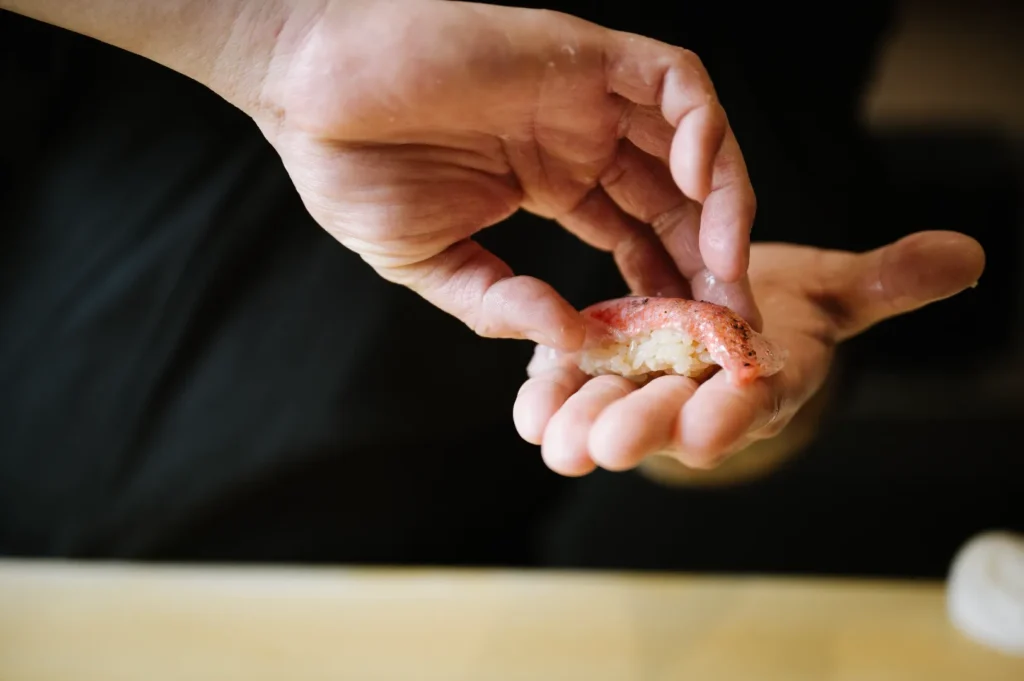 Chef Masa holding a piece of sushi with delicate care, showcasing a slice of pink fish atop white rice. The scene conveys precision and culinary skill.