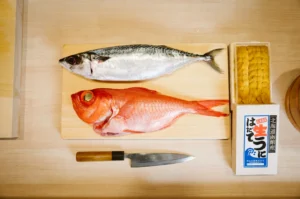 Fresh fish on a wooden board: a shiny mackerel and a vibrant red snapper. A sharp knife and a box of yellow sea urchin roe are nearby.