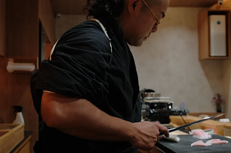 Chef Masa in a dimly lit kitchen meticulously slices pink fish fillets on a black cutting board, conveying focus and precision. The warm tones create an intimate atmosphere.