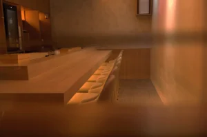 Warmly lit, intimate bar with a long wooden counter, rows of beige stools neatly aligned underneath, and minimalist decor creating a cozy atmosphere.