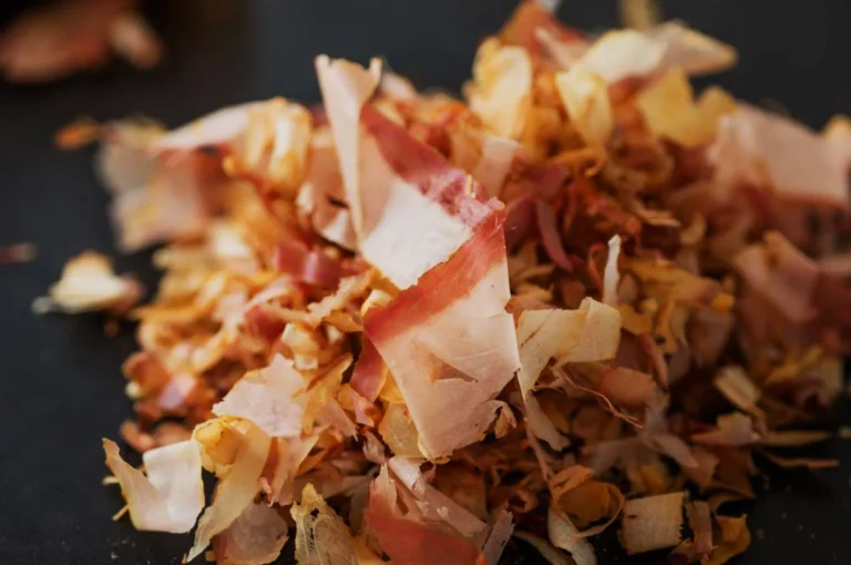 A close-up of a pile of thin, light brown and pinkish bonito flakes on a dark surface, conveying a sense of delicacy and umami flavor.
