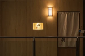 Dimly lit Japanese restaurant entrance with a beige curtain door, softly glowing wall light, and illuminated sign featuring Japanese characters. Cozy ambiance.