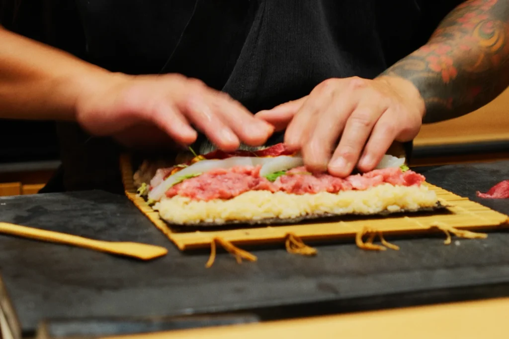 Hands skillfully preparing sushi, pressing ingredients on a bamboo mat. Rice, thinly sliced vegetables, and raw meat are visible. A focused, artisanal vibe.