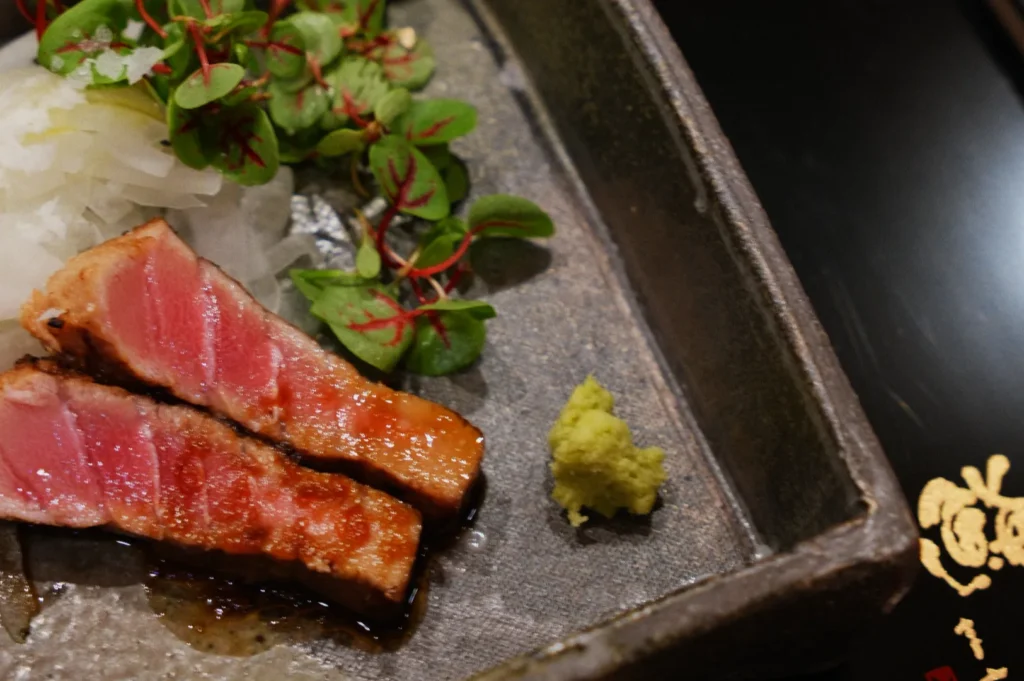 Sliced seared tuna with pink center on a textured plate, garnished with grated radish, microgreens, and a dollop of green wasabi, creating a fresh, elegant presentation.