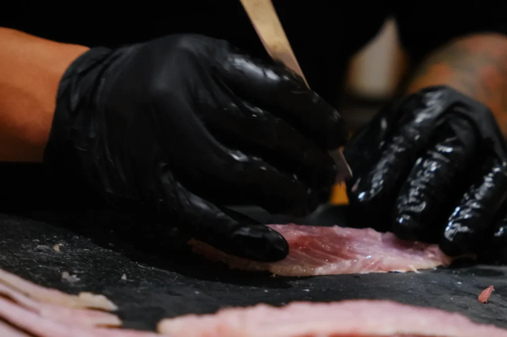 Hands in black gloves carefully slice raw fish on a dark cutting board, emphasizing precision and concentration in food preparation.