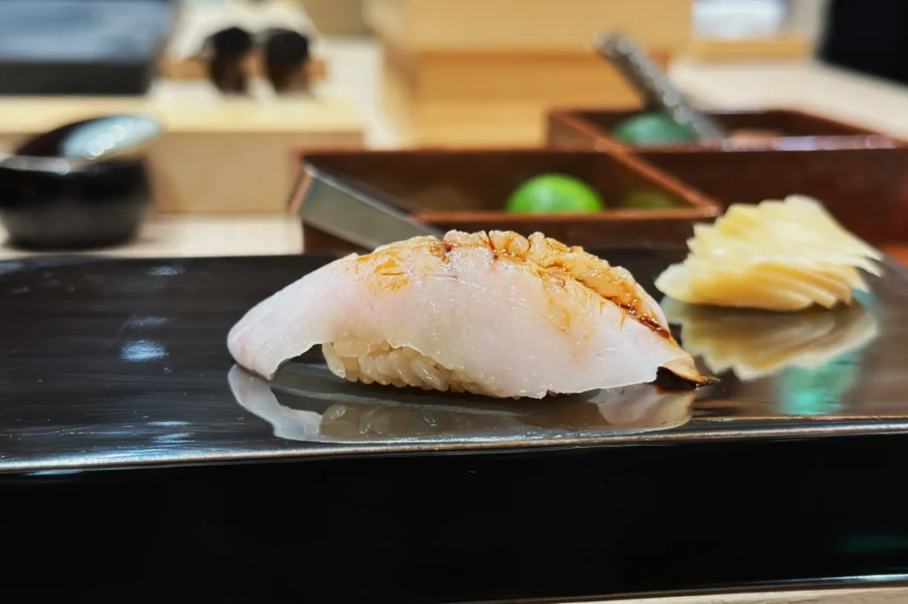 Close-up of a sushi piece on a glossy black plate, featuring white fish with a seared top and ginger slices beside it, set in a refined, minimalistic setting.