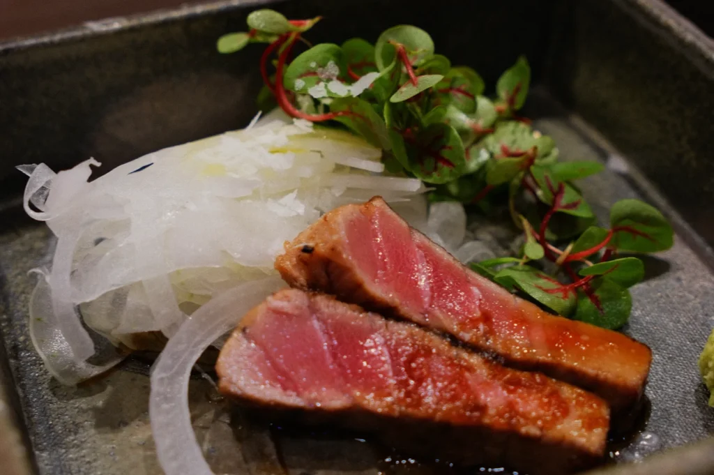 Seared tuna slices with a pink center on a plate, accompanied by thinly sliced white onions and fresh green salad leaves, creating a fresh and elegant presentation.