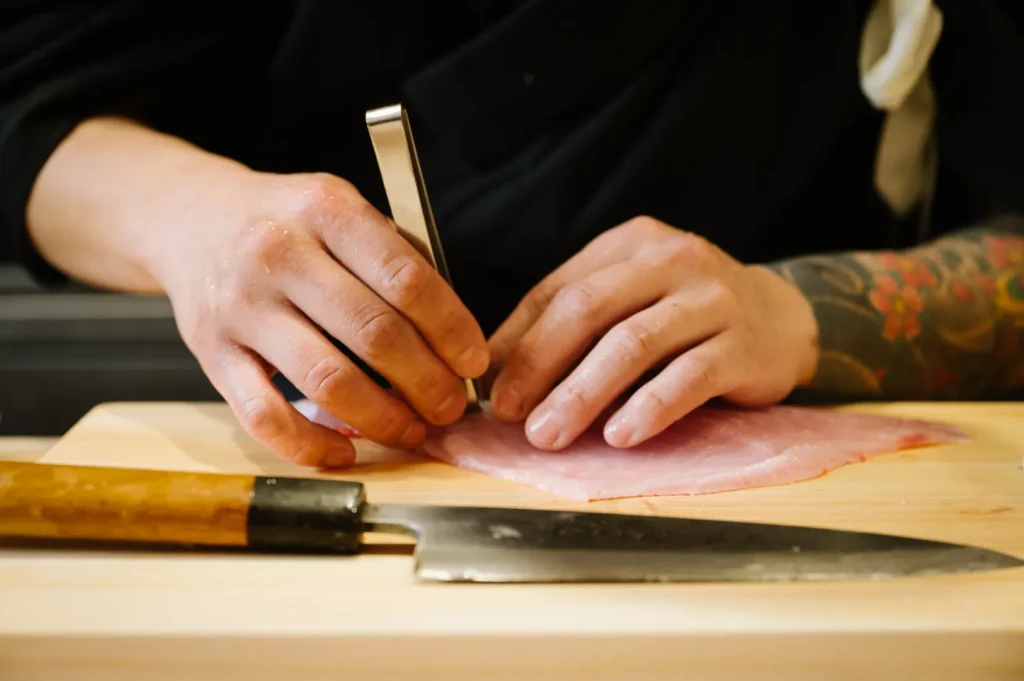 Close-up of chef Masa with tattooed arms using tweezers to prepare raw fish on a wooden board, a sharp knife nearby, conveying precision and focus.