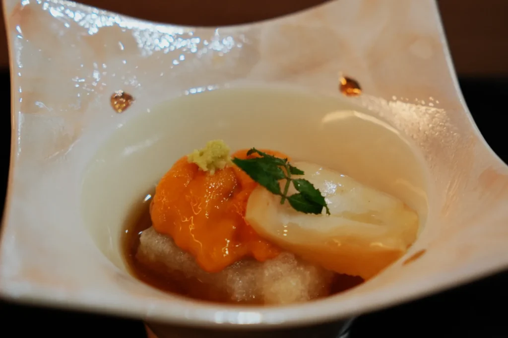 A delicate dish in a white, intricately designed bowl, featuring creamy sea urchin, wasabi, and garnished with a green herb, conveying an elegant and refined presentation.