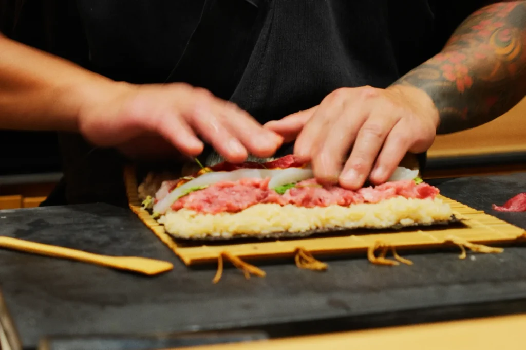Chef Masa skillfully prepare sushi on a bamboo mat, layering rice, vibrant pink fish, and vegetables. The mood is focused and meticulous, emphasizing craftsmanship.