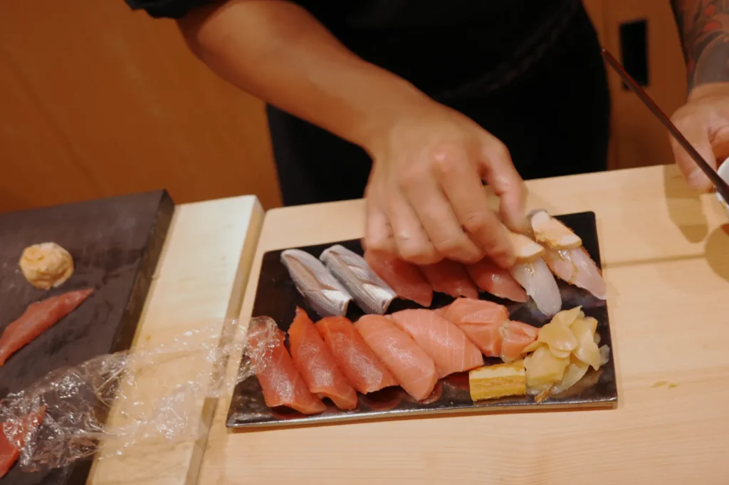 Hands arranging assorted sushi on a black plate, featuring various types of fish and garnishes. The setting conveys skill and attention to detail.