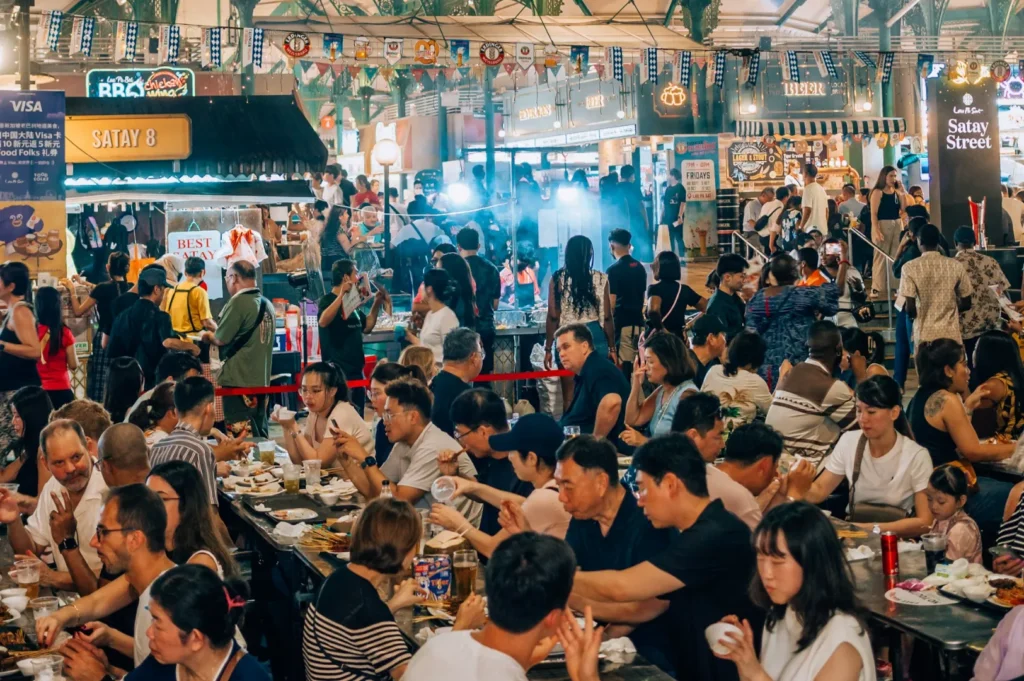A bustling night market with diverse food stalls, colorful banners, and a large crowd enjoying meals, creating a lively and vibrant atmosphere.