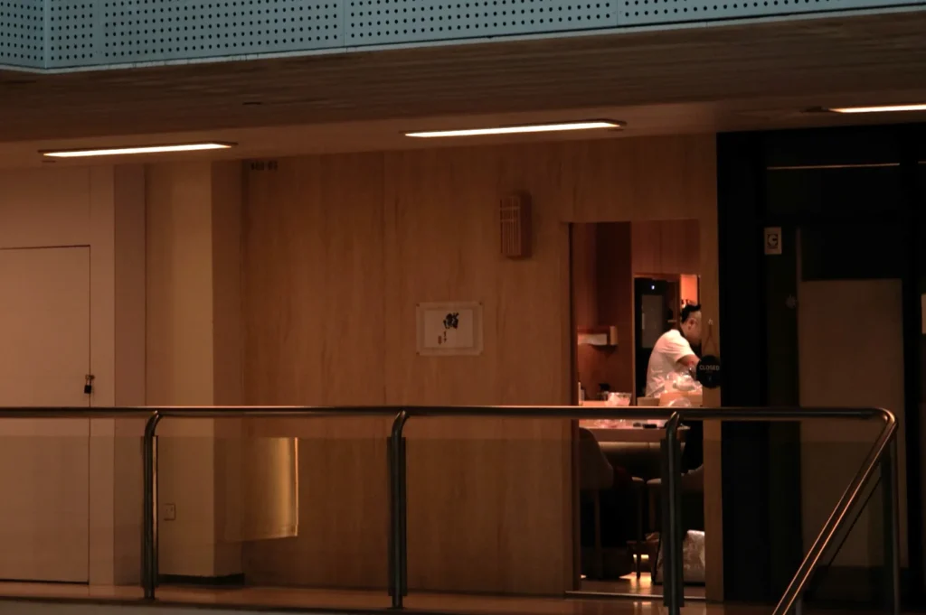 A dimly lit interior with Chef Masa in a white shirt inside a room, seen through a small window. The scene feels calm and secluded, framed by modern architecture.