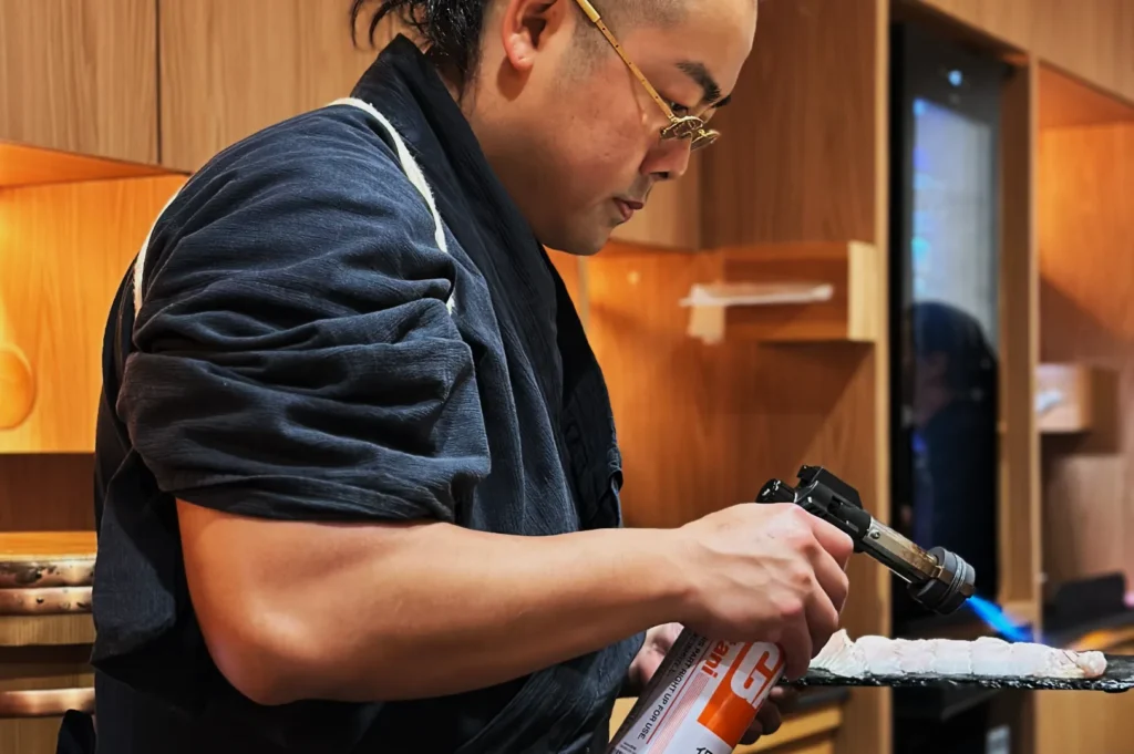 Chef Masa in a dark uniform intensively torches food with a blowtorch in a modern kitchen. The scene conveys focus and culinary precision.