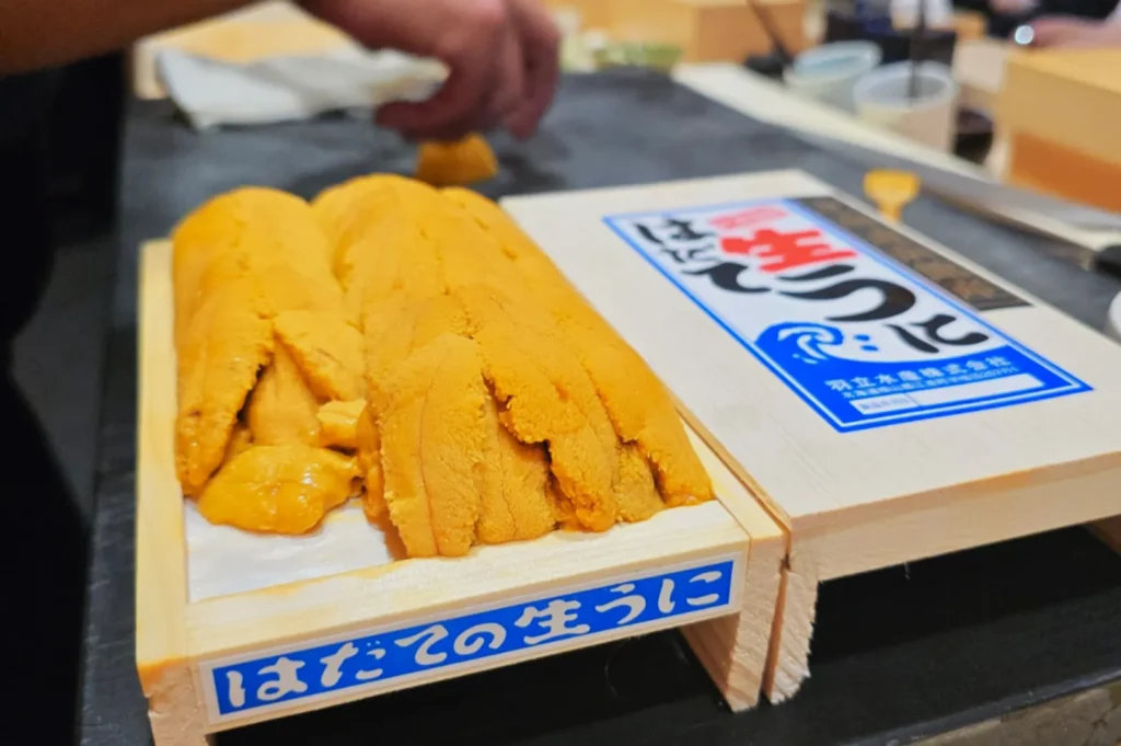 Fresh sea urchin (uni) is displayed in a wooden box on a black counter. A hand is seen in the background. The scene conveys freshness and craftsmanship.