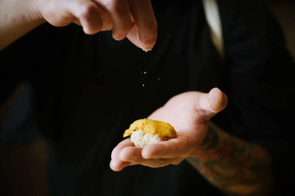 Chef Masa delicately sprinkles salt onto a piece of sushi held in their tattooed hand. The sushi rests on rice, topped with a yellow uni, creating a focused, artistic culinary moment.