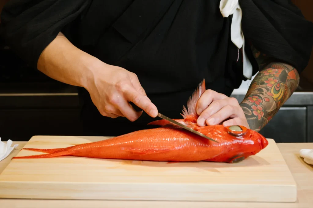 Chef Masa in a black uniform, with a tattooed arm, skillfully fillets a vibrant red fish on a wooden cutting board, conveying precision and focus.