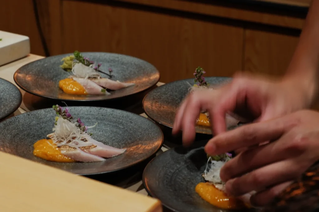 Chef garnishing plates of sashimi with herbs and orange sauce. The dark dishes contrast with the delicate fish, creating an elegant presentation.