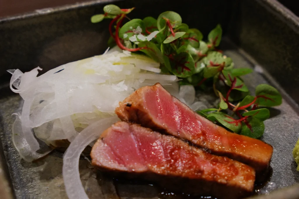 Seared tuna slices with a pink center are arranged on a plate. They are accompanied by sliced onions and fresh greens, creating an appetizing presentation.