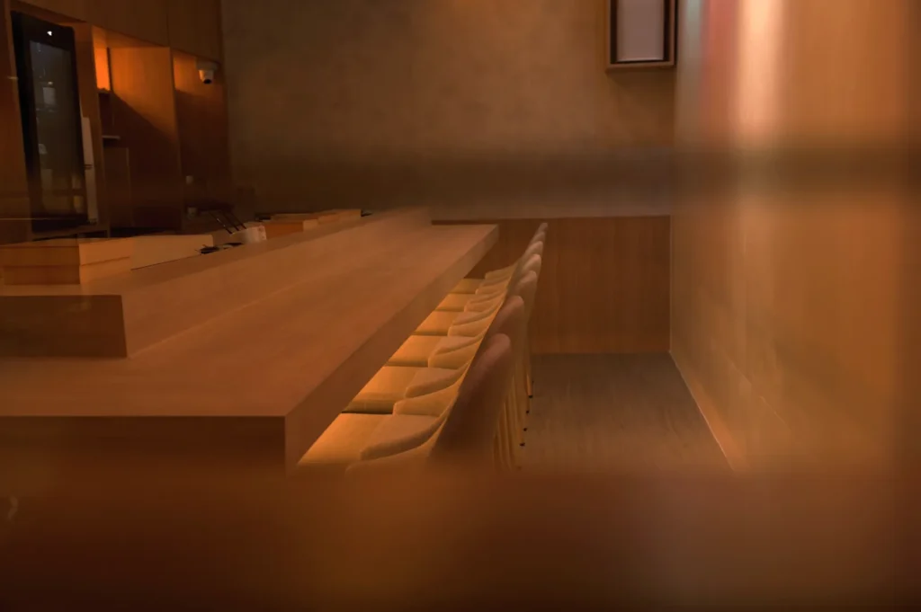 Warmly lit, minimalist interior with a long wooden bar counter and neatly arranged stools, creating a cozy, inviting ambiance.