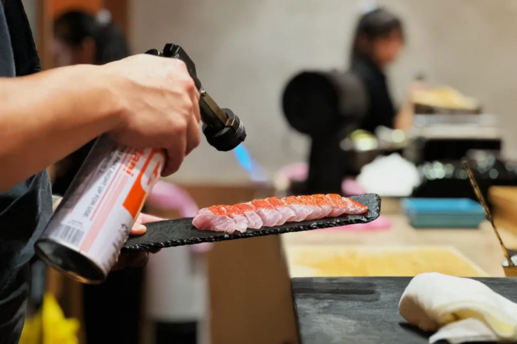 Chef uses a torch to sear slices of fish on a black slate in a sushi restaurant. The atmosphere is lively, with blurred staff in the background.
