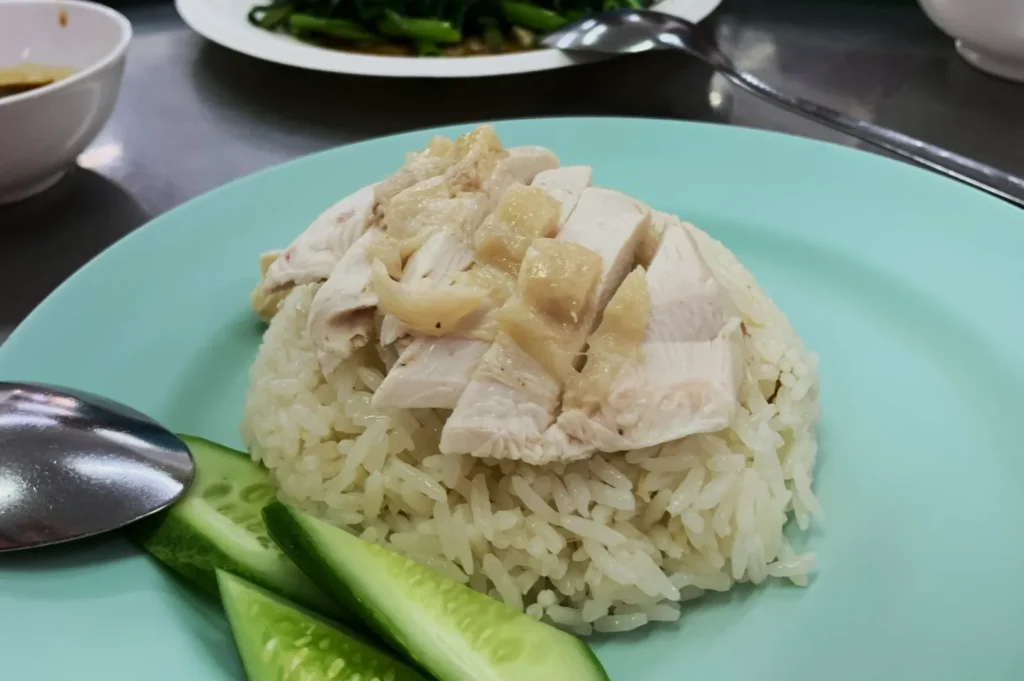 Chicken and rice on a light blue plate, garnished with sliced cucumber. Served with a spoon; background shows green vegetables and a bowl.
