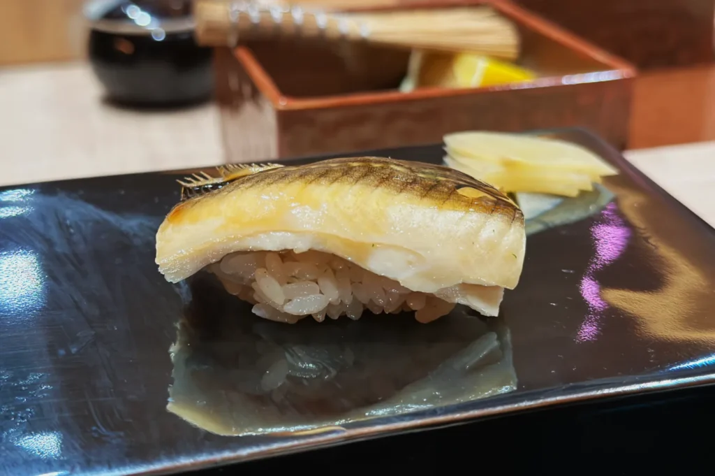 A piece of mackerel nigiri sushi rests on a black plate, with glossy rice underneath. Pickled ginger is placed beside it, creating a fresh and elegant presentation.