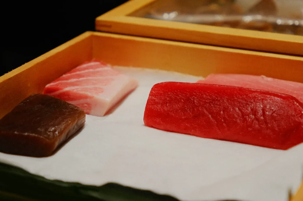 A wooden tray holds slices of fresh fish. From left to right: a dark piece, a marbled pink piece, and a bright red piece, all placed on white paper.