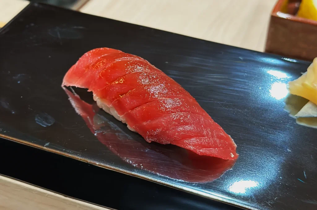 Tuna nigiri sushi on a glossy black plate, showcasing a slice of vibrant red tuna atop rice. The setting is elegant and minimalistic.