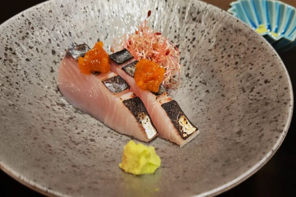 Sliced seared fish with charred skin and orange garnish on a speckled gray plate, accompanied by a small wasabi mound and sprouts, evoking elegance.