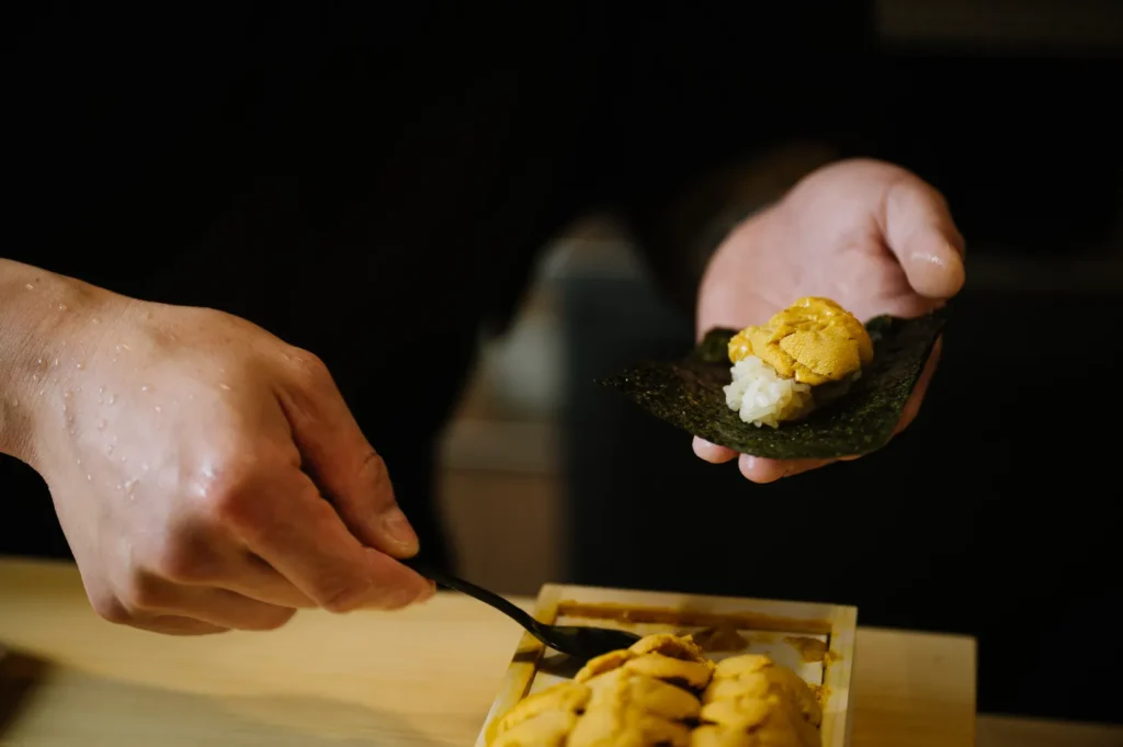 Close-up of hands of Chef Masa preparing sushi with sea urchin, rice, and nori. A spoon adds uni atop the rice, conveying precision and culinary artistry.