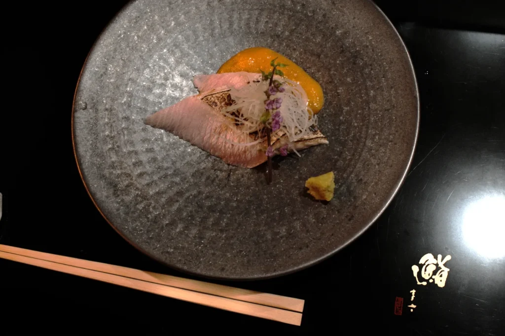 A delicate plate of sashimi with thinly sliced fish, garnished with purple flowers, radish, and a dollop of sauce, creating an elegant and refined presentation. Chopsticks rest beside the dark, textured plate.