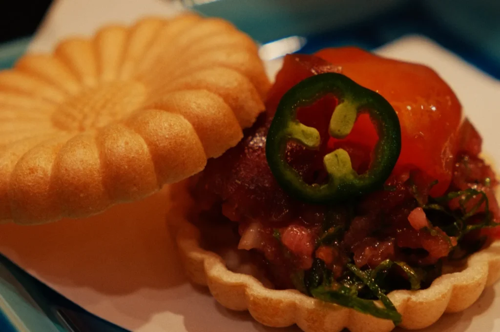 A delicate open-faced tart with a scalloped edge filled with diced raw fish, topped with a slice of jalapeño and a dollop of orange jelly, resting on a blue plate.