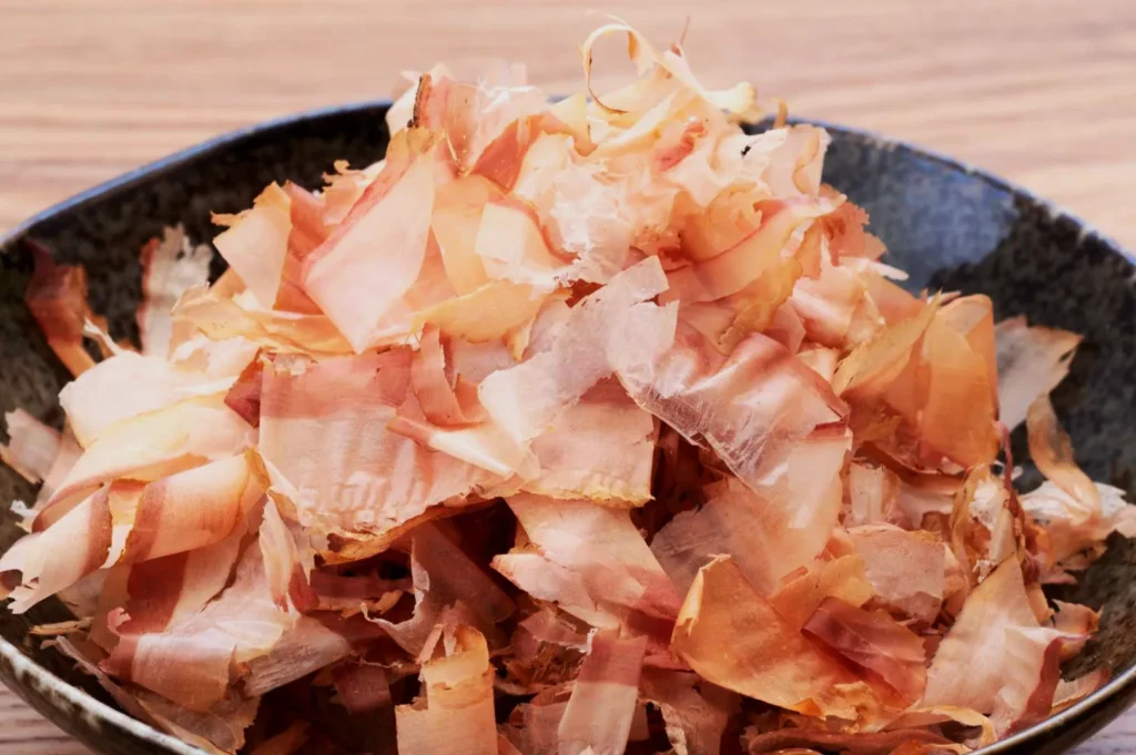 A dark bowl filled with thin, translucent, pale pink bonito flakes sits on a wooden surface. The delicate flakes appear light and airy.