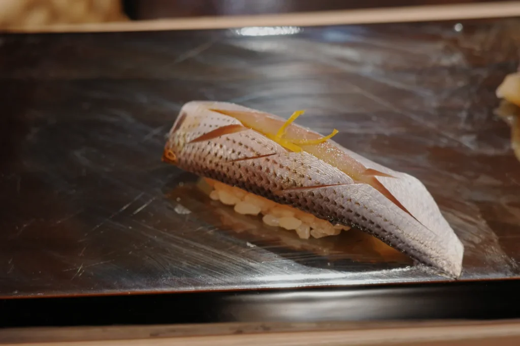 Close-up of a piece of sushi featuring shiny silver fish with scored skin, garnished with a small yellow strip, set on rice atop a dark plate.