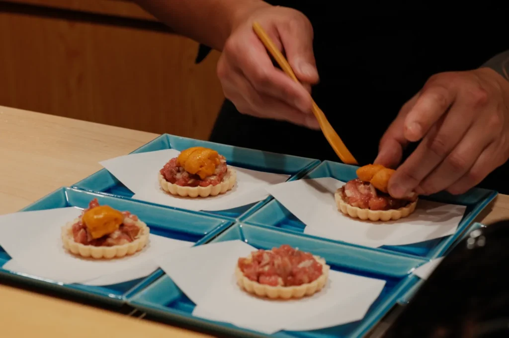 Hands using chopsticks to place uni on tartlets with raw fish, arranged on square blue plates lined with paper, conveying a delicate culinary preparation.
